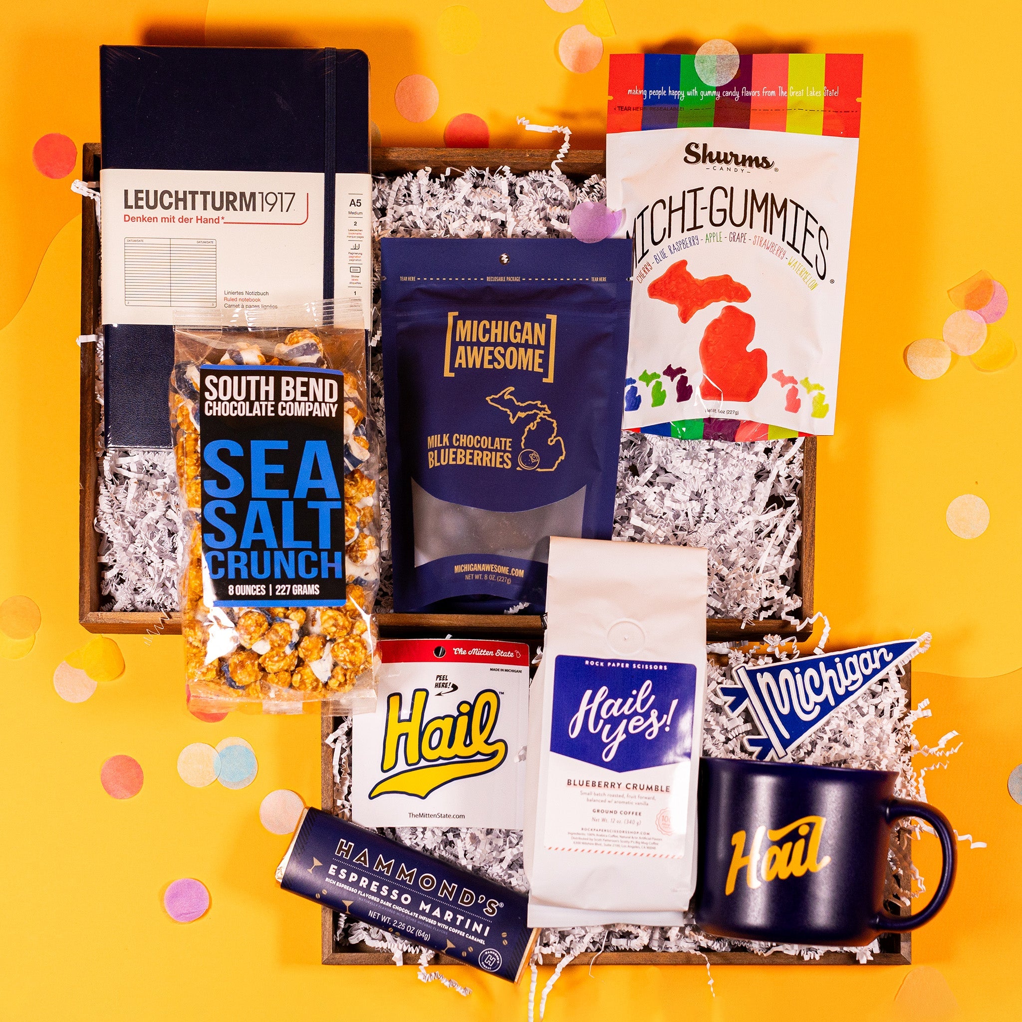 On a bright yellow background with colorful confetti sits a collection of items in a gift box with white crinkle paper. A navy blue notebook, a clear bag of Sea Salt Crunch Popcorn, a navy blue pouch of Milk Chocolate Blueberries, a pouch of Michi-gummies, a white bag of Blueberry Crumble coffee, a small pennant sticker that says Michigan, a navy blue mug that says "Hail", a sticker of the word "Hail" in yellow script, and an Espresso Dark Chocolate Bar with a navy blue wrapper and gold foil.