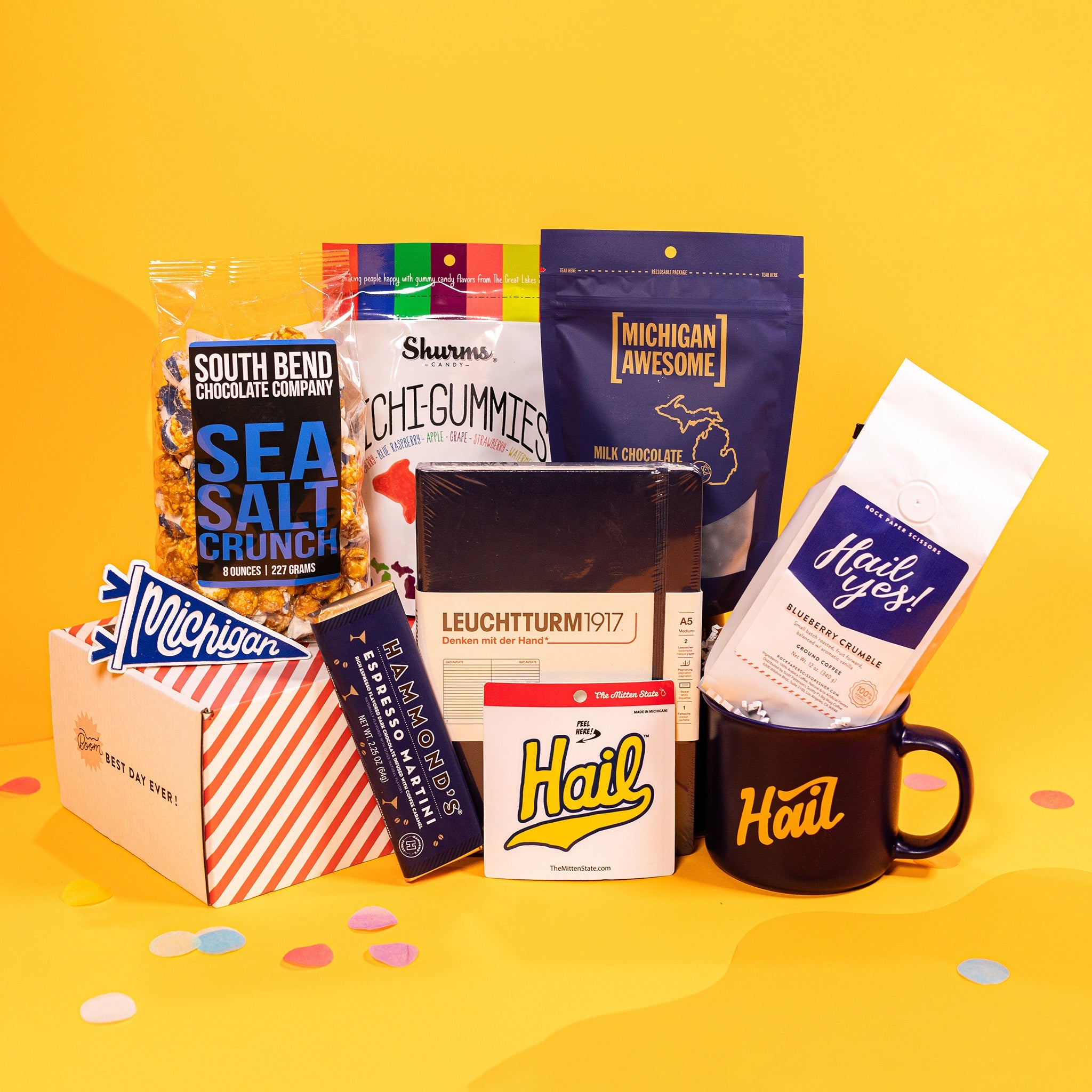 On a bright yellow background with colorful confetti sits a collection of items in a gift box with white crinkle paper. A navy blue notebook, a clear bag of Sea Salt Crunch Popcorn, a navy blue pouch of Milk Chocolate Blueberries, a pouch of Michi-gummies, a white bag of Blueberry Crumble coffee, a small pennant sticker that says Michigan, a navy blue mug that says "Hail", a sticker of the word "Hail" in yellow script, and an Espresso Dark Chocolate Bar with a navy blue wrapper and gold foil.