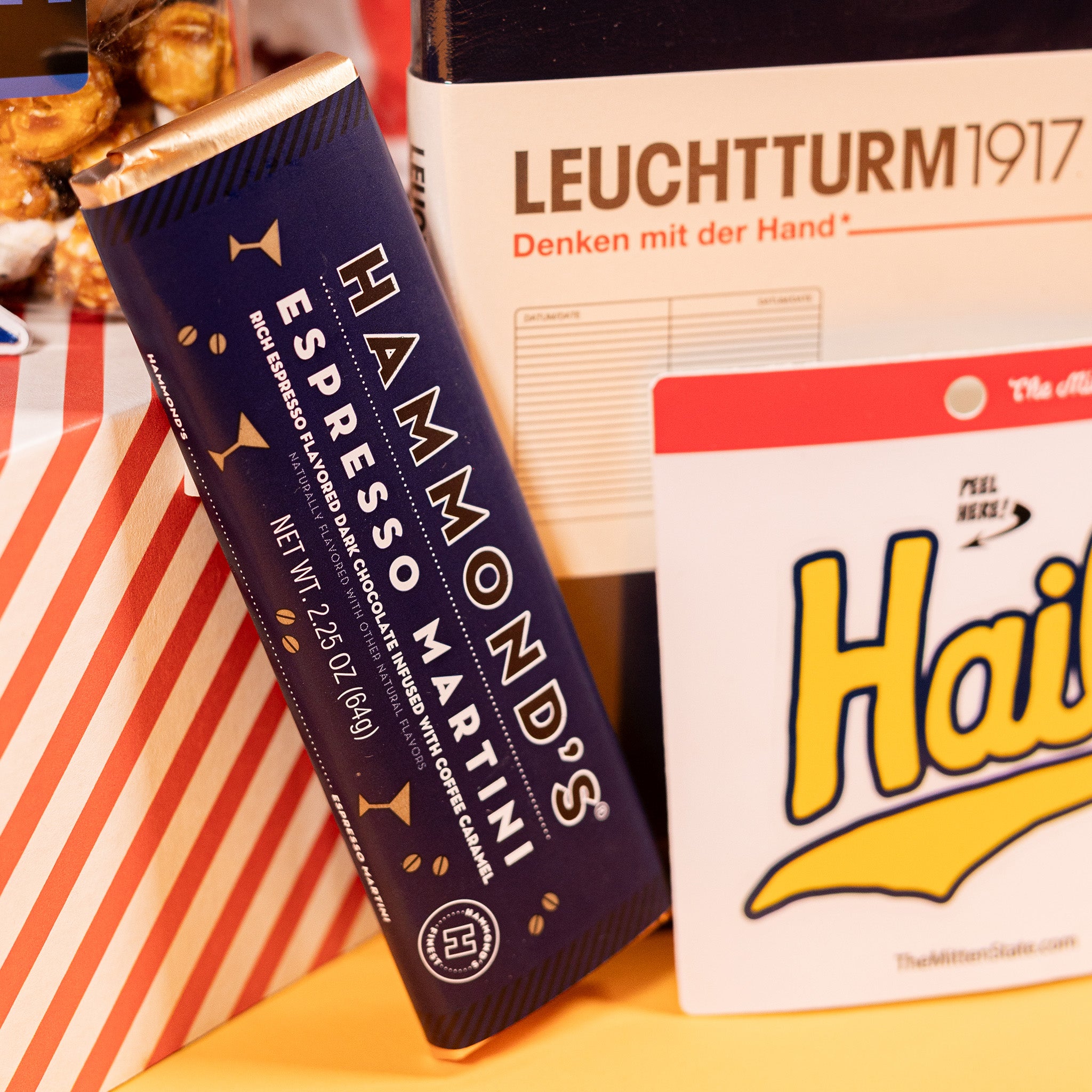 On a bright yellow background with colorful confetti sits a collection of items next to a red striped gift box: an A5 size navy blue notebook from Leuchtturm1917, a die-cut sticker of the word "Hail" in yellow script, and a 2.25 oz Espresso Martini Dark Chocolate Bar with a navy blue wrapper and gold foil from Hammond's.