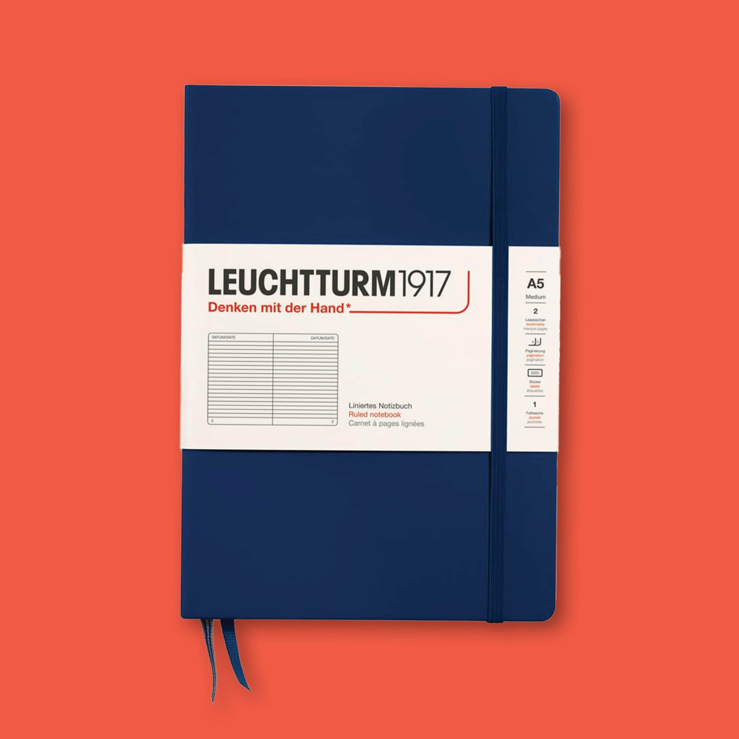 On a tomato red background sits an A5 size navy blue notebook from Leuchtturm1917 with navy blue elastic closure and navy blue bookmark ribbons.