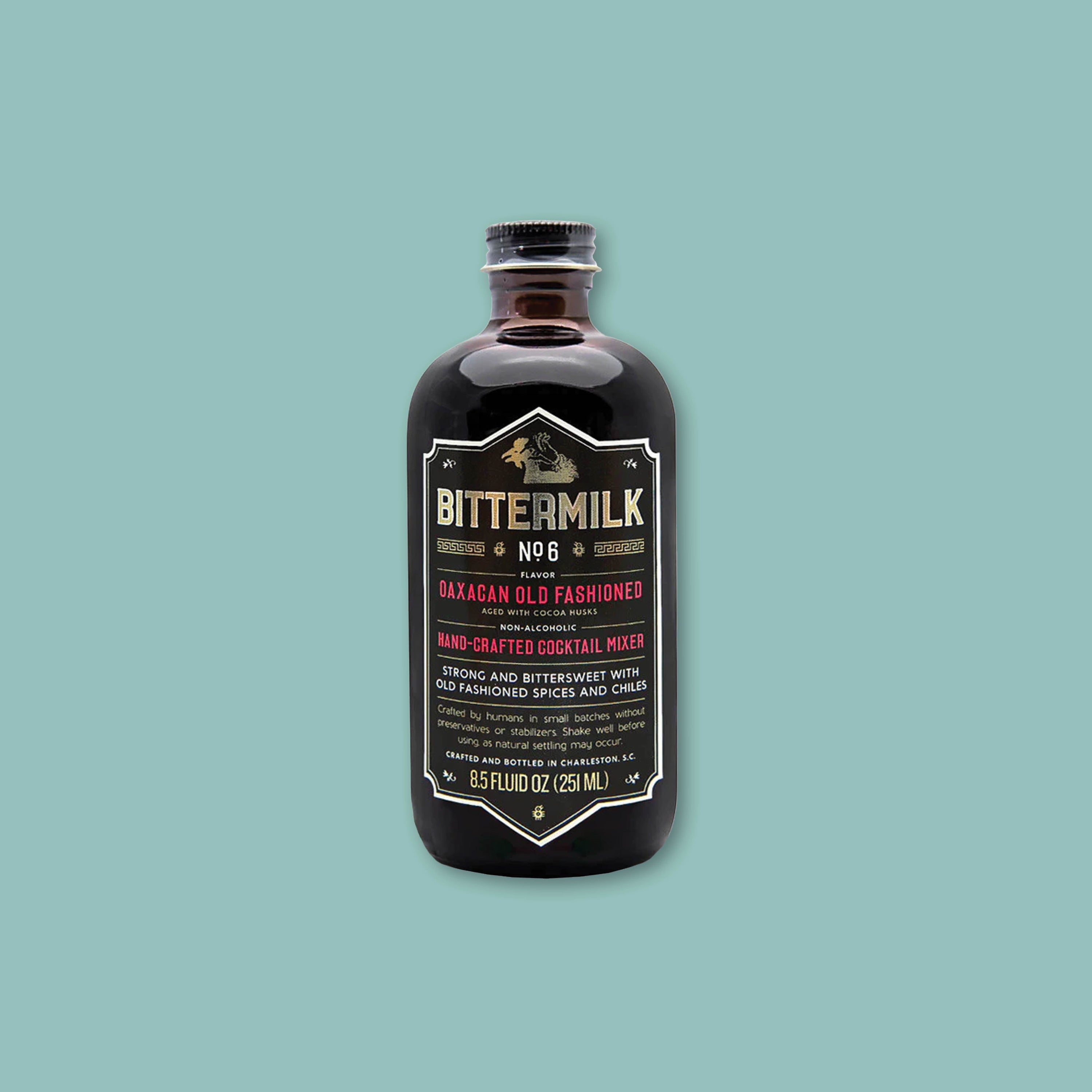 On a teal background sits a dark brown bottle of Bittermilk No. 6 flavor Oaxacan Old Fashioned Handcrafted Cocktail Mixer.