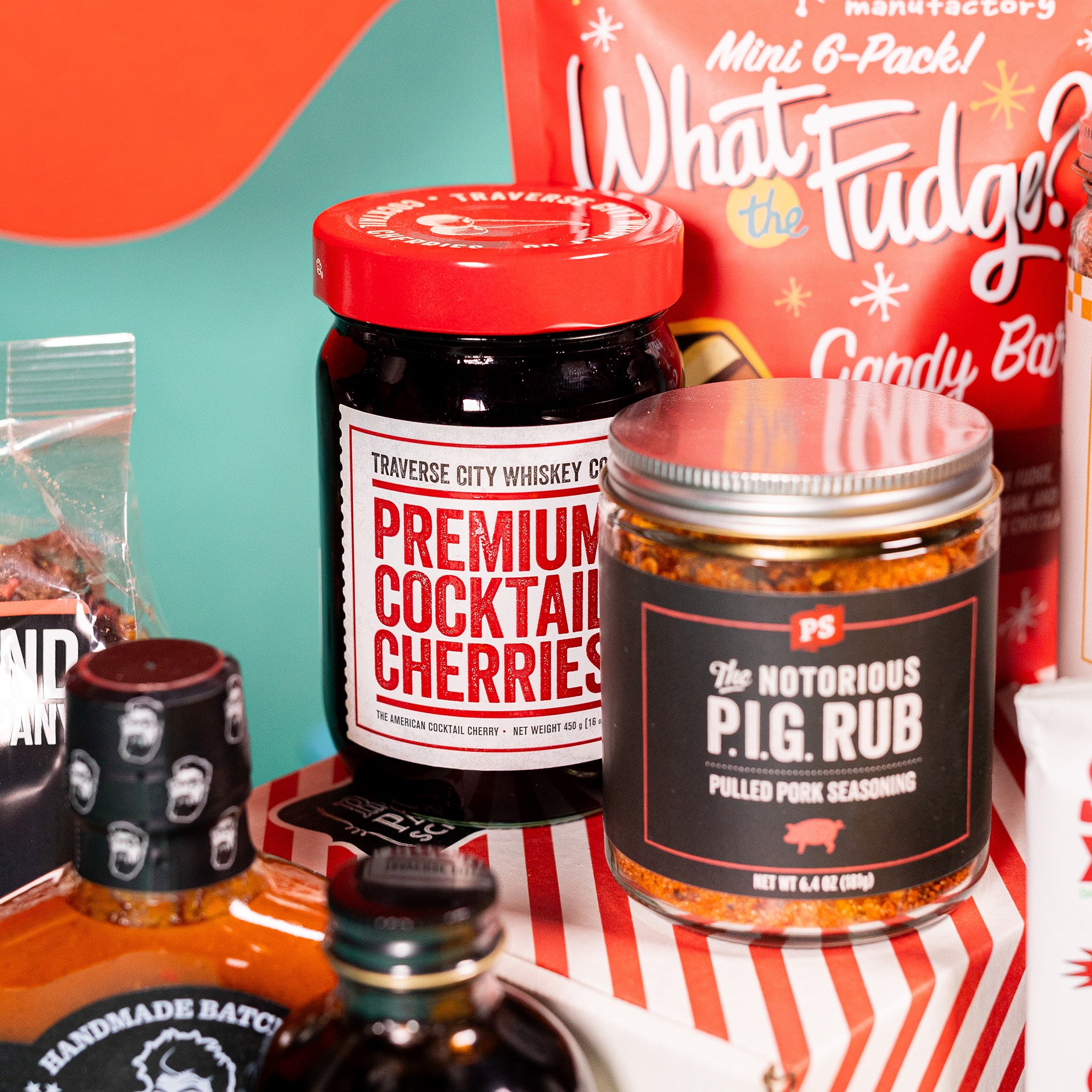 On a red and teal background sits a red bag of Zingerman's candy bars, a jar of The Notorious P.I.G. Run, and a jar of Premium Cocktail Cherries with a red lid.