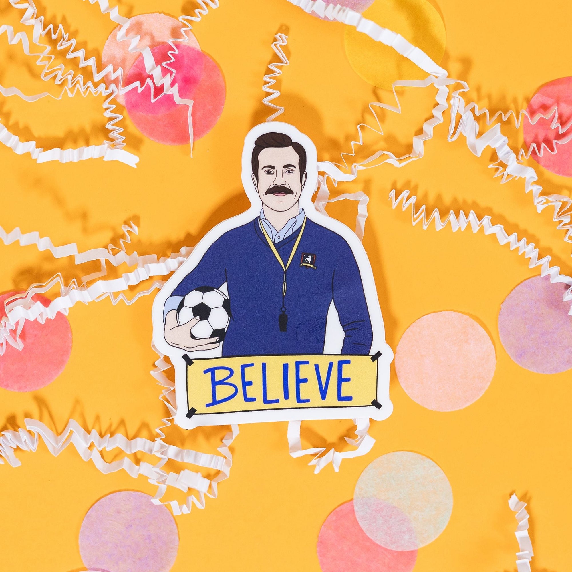 On a sunny mustard background sits a sticker with white crinkle and big, colorful confetti scattered around. This sticker is an illustration of Ted Lasso holding a futbol with a sign at the bottom that says "BELIEVE" in blue handwritten lettering. The colors are in navy, yellow, blue, and light blue with black and white. 2"-3"