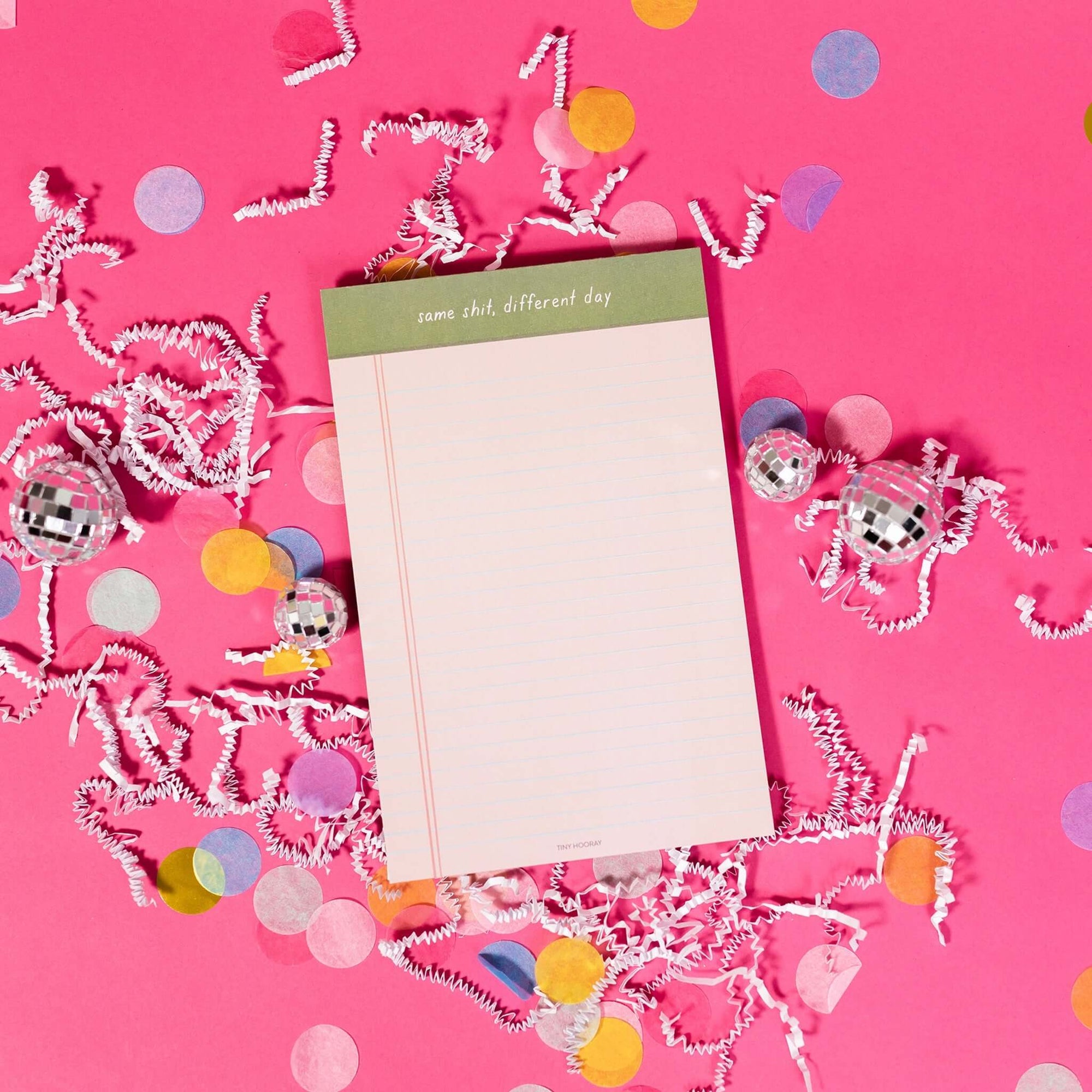 On a hot pink background sits a light pink notepad with white crinkle and big, colorful confetti scattered around. There are mini disco balls. It says "same shit, different day" at the top and has blue lines.