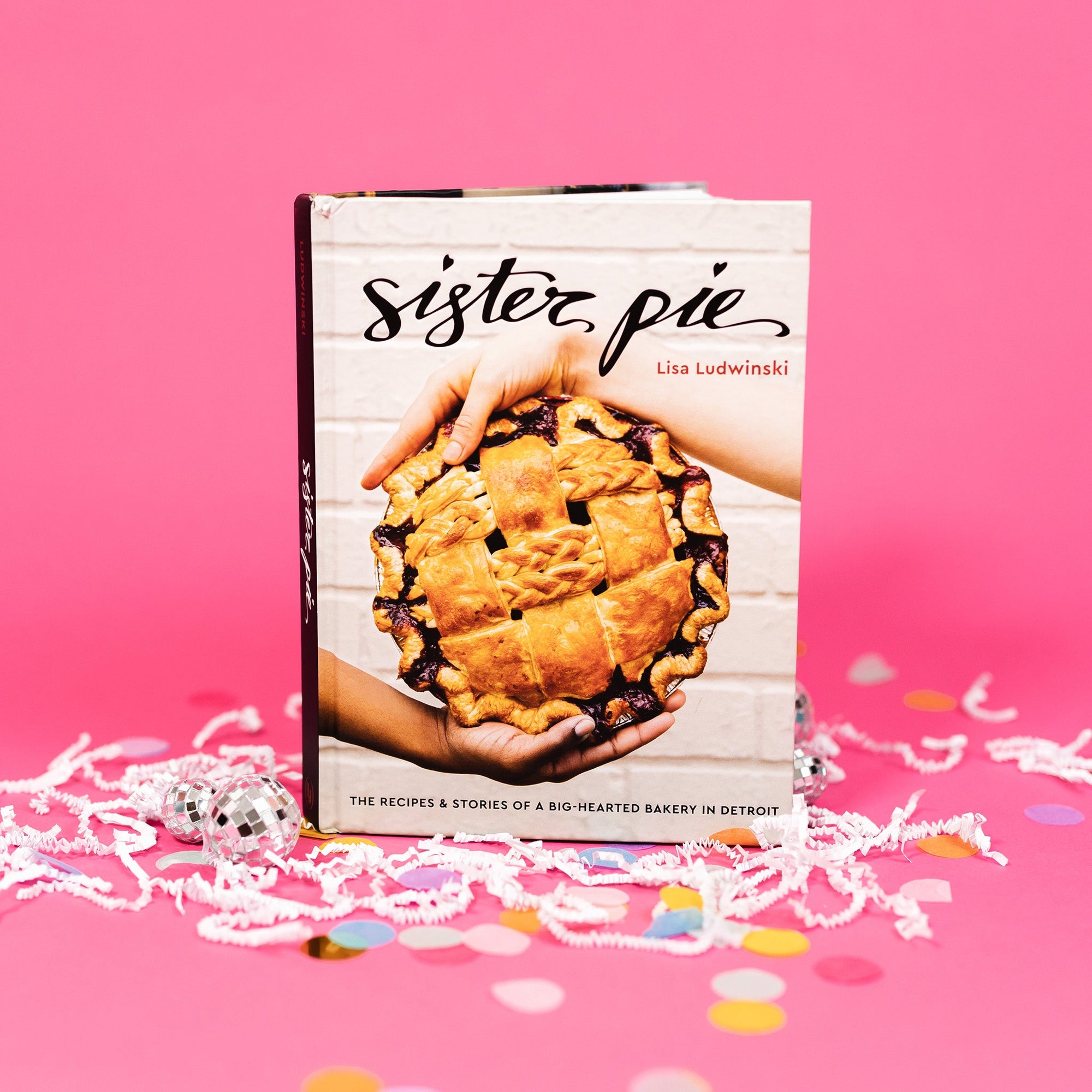 On a hot pink background sits a book with white crinkle and big, colorful confetti scattered around. There are mini disco balls. This cookbook says "sister pie" in a black handwritten lettering font and has a pitcure of two hands holding a beautiful pie with a white, brick background. On the bottom is says "The recipes & stories of a big-hearted bakery in Detroit" in a black, all caps block font.