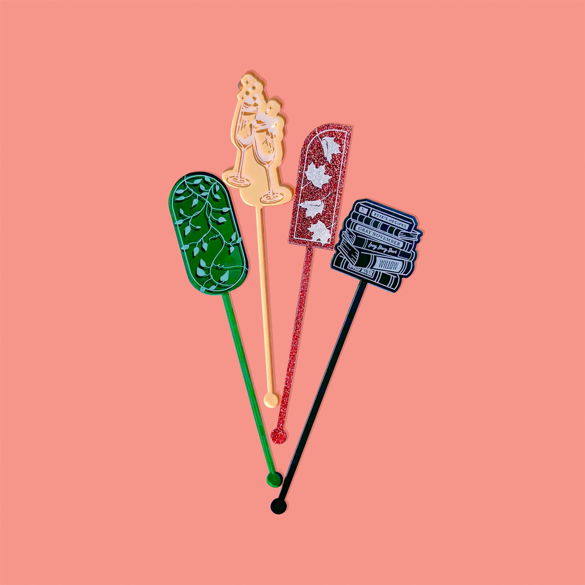 On a coral pink background sits acrylic stirrers. This Taylor Swift inspired set of 4 stirrers includes a dark green transparent ivy, a gold transparent bubbling champagne glasses, copper glitter fall leaves, and a bblack smoke book stack.