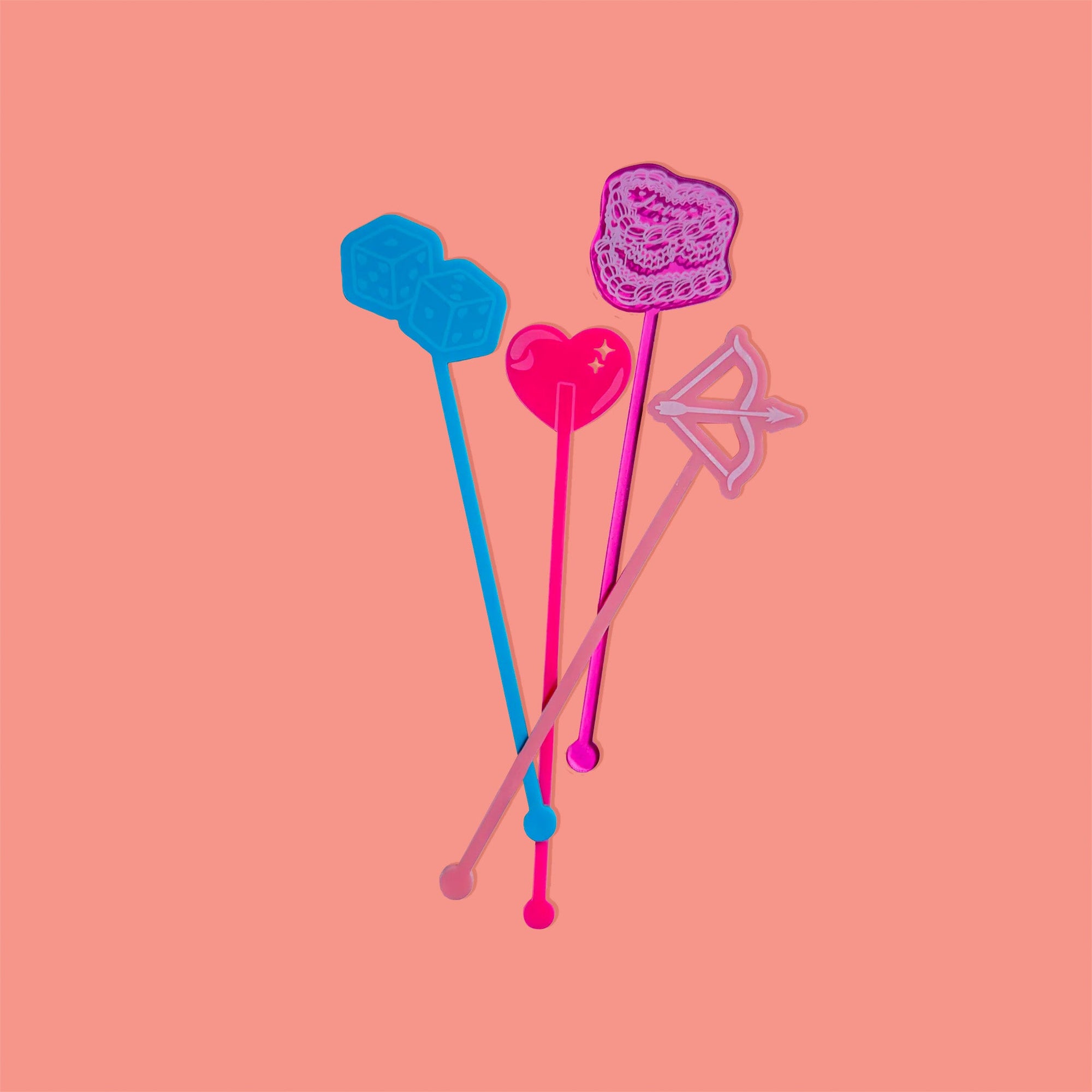 On a coral pink background sits acrylic stirrers. This Taylor Swift inspired set of 4 stirrers includes a pair of laguna opaque heart dice, a pink opague heart lollipop, a salmon opaque bow and arrow, and a pink transparent Lover cake.