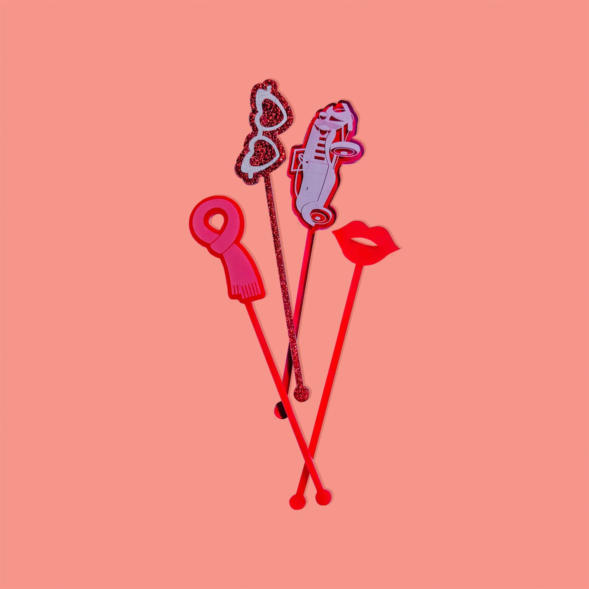 On a coral pink background sits acrylic stirrers. This Taylor Swift inspired set of 4 stirrers includes a red opaque scarf, red glitter heart sunglasses, a red transparent vintage car, and red translucent lips.