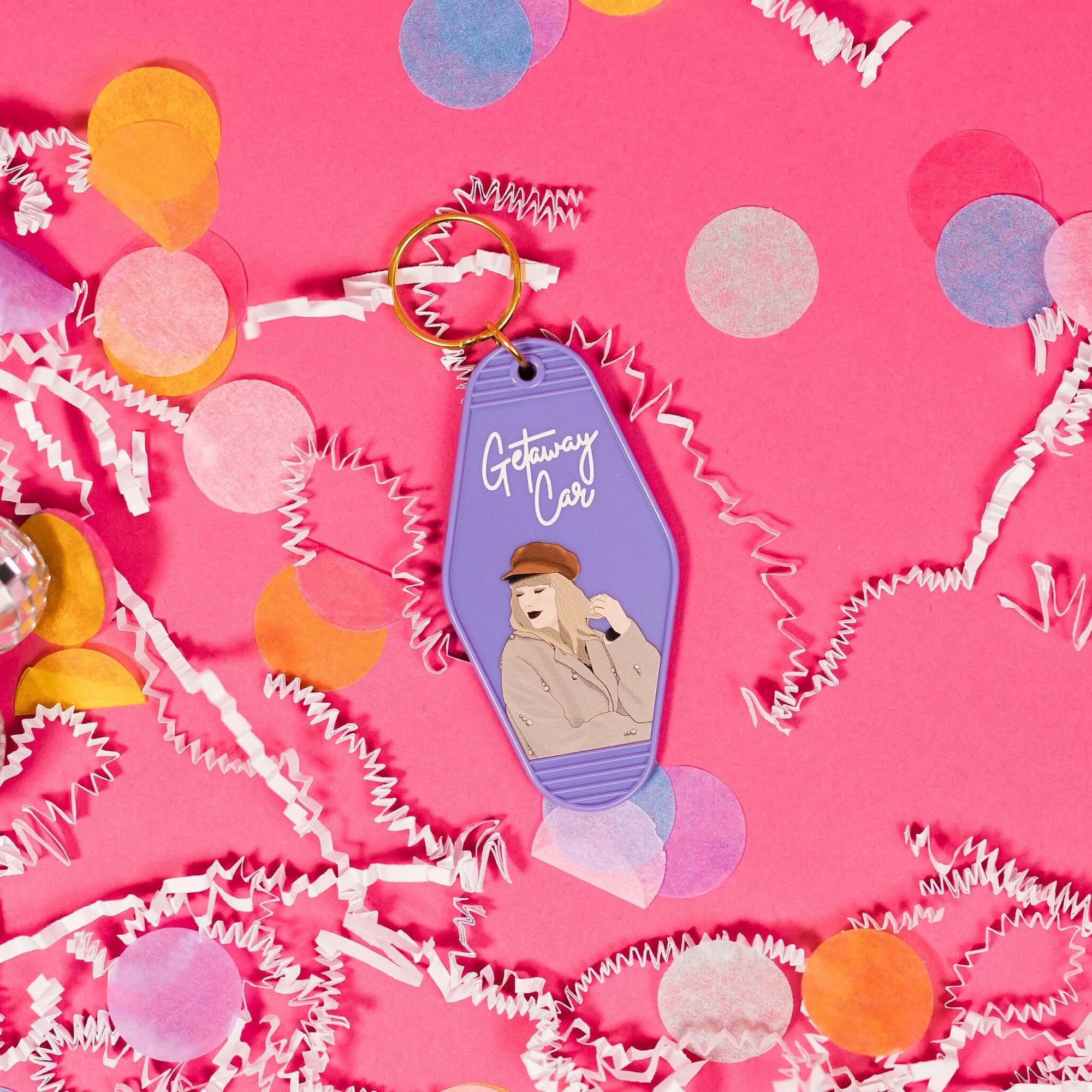 On a hot pink background sits a keychain with white crinkle and big, colorful confetti scattered around. There are mini disco balls. This Taylor Swift inspired vintage, motel keychain is lavender with an illustration of Taylor Swift wearing a hat and coat. In a white, handwritten lettering script it says "Getaway Car".