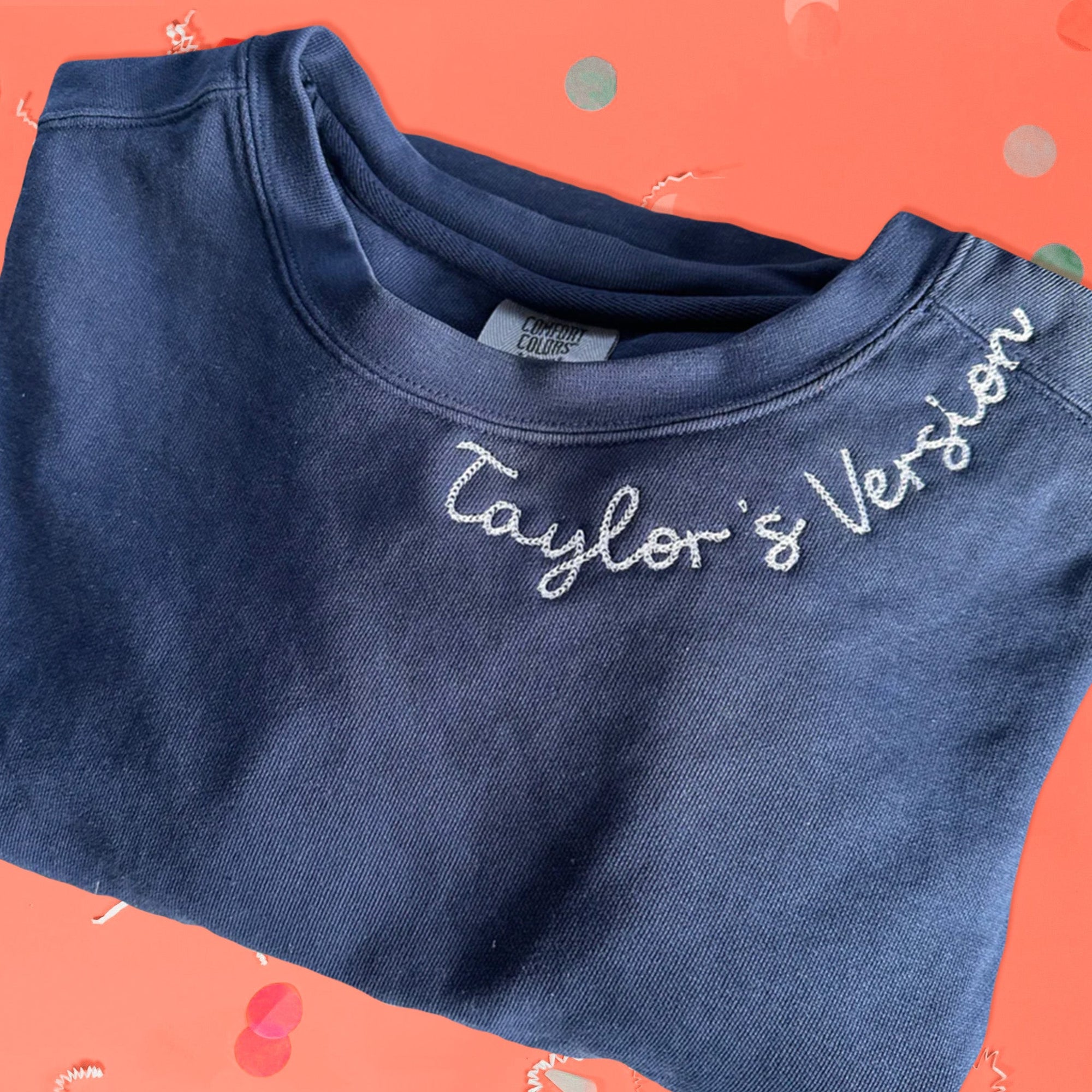 On a pink coral background sits the front of a sweatshirt with white crinkle and big, colorful confetti scattered around. This Taylor Swift Inspired crewneck navy sweatshirt has white chain stitched script lettering around the neck and it says "Taylor's Version."