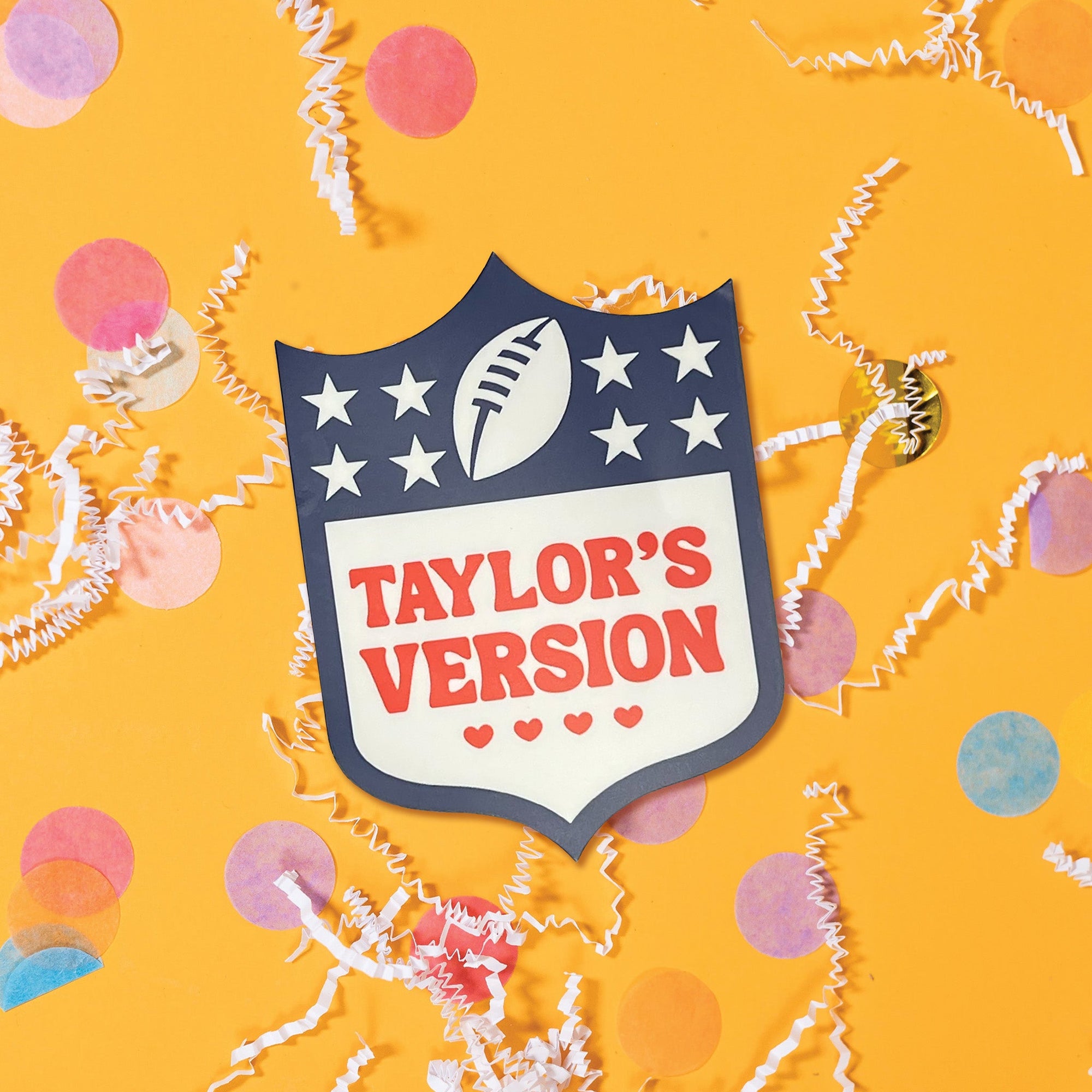 On a hot pink background sits a mug on top of a red and white striped packing tape with white crinkle and big, colorful confetti scattered around. There are mini disco balls. This NFL-Taylor Swift inspired mug has an NFL logo on the front and it says "TAYLOR'S VERSION" with four hearts under it. It is in red.