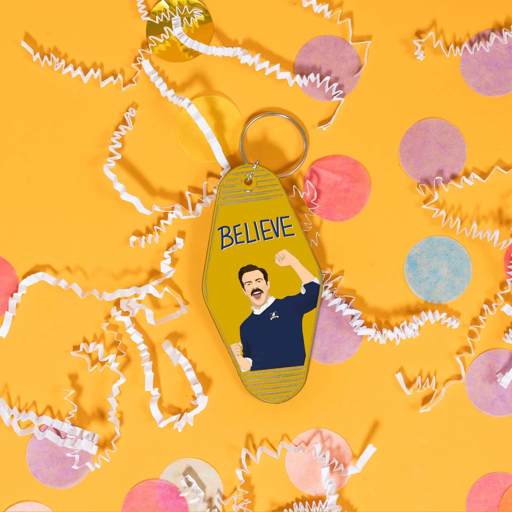 On a sunny mustard background sits a keychain with white crinkle and big, colorful confetti scattered around. This Ted Lasso inspired vintage, motel keychain is golden yellow with an illustration of Ted Lasso and it says "BELIEVE" in a navy, handwritten lettering block font.