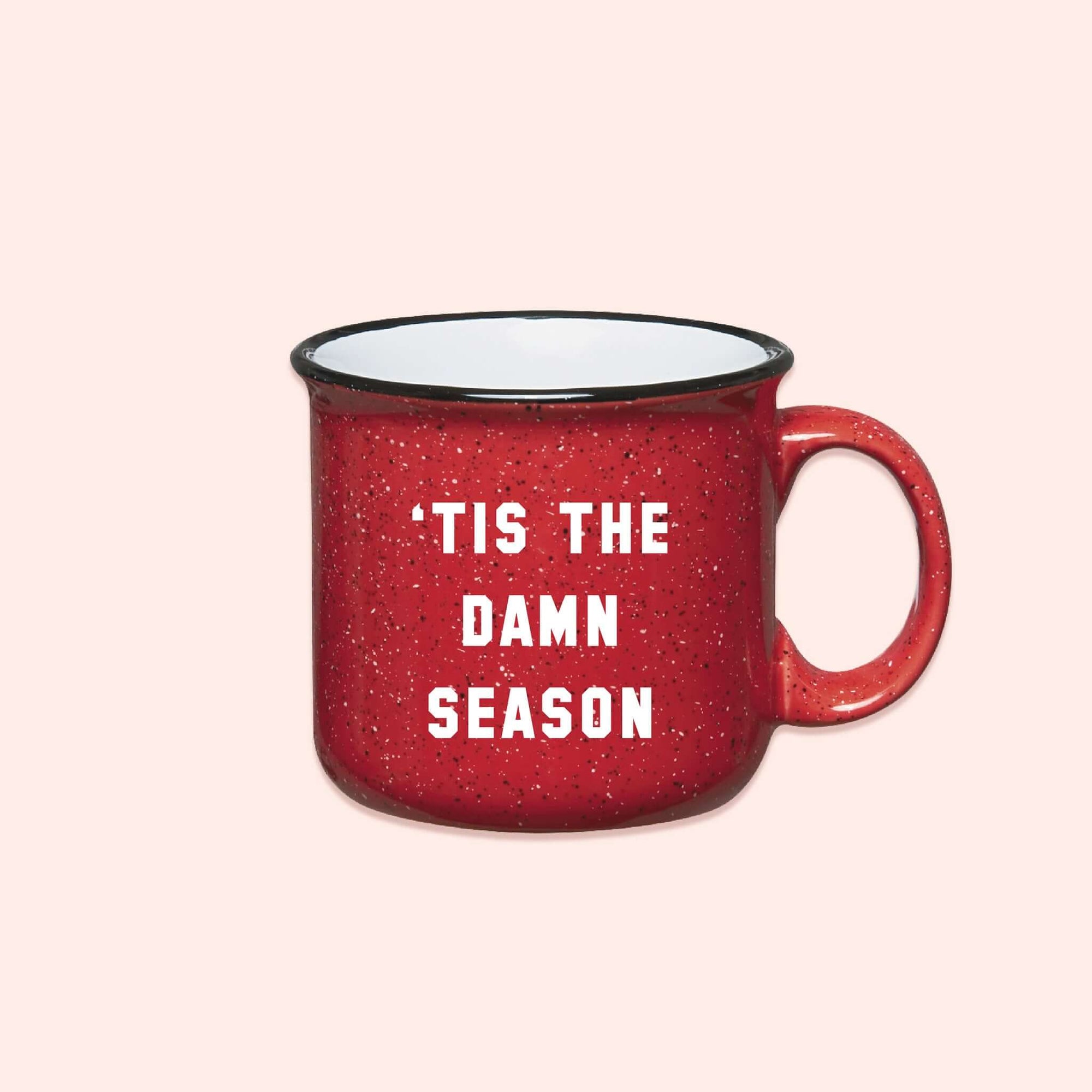 On a coral pink background sits a red campfire mug with black and white specks. It says "'TIS THE DAMN SEASON" in white collegiate lettering.