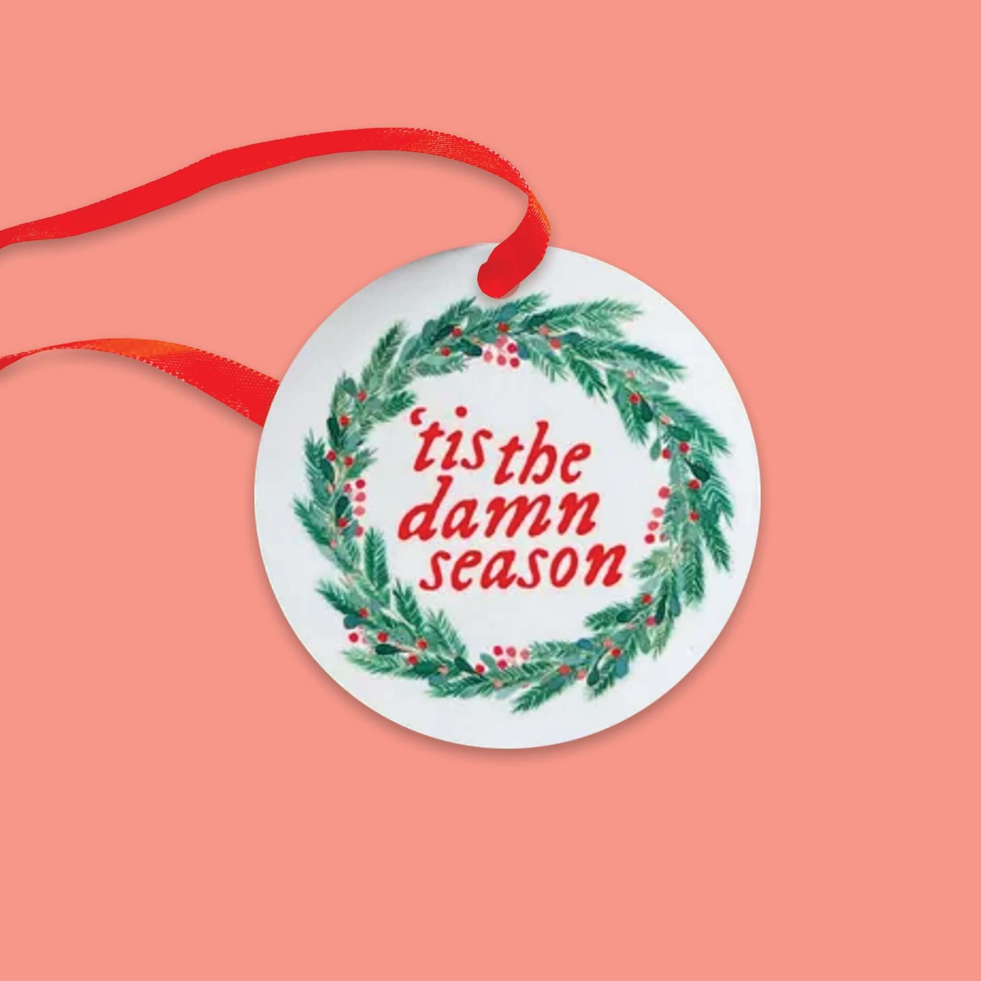 On a coral pink background sits a round ornament. This Taylor Swift inspired ceramic ornament is white with an illustration of a wreath with red berries. It says "'tis the damn season" in thick red lettering.