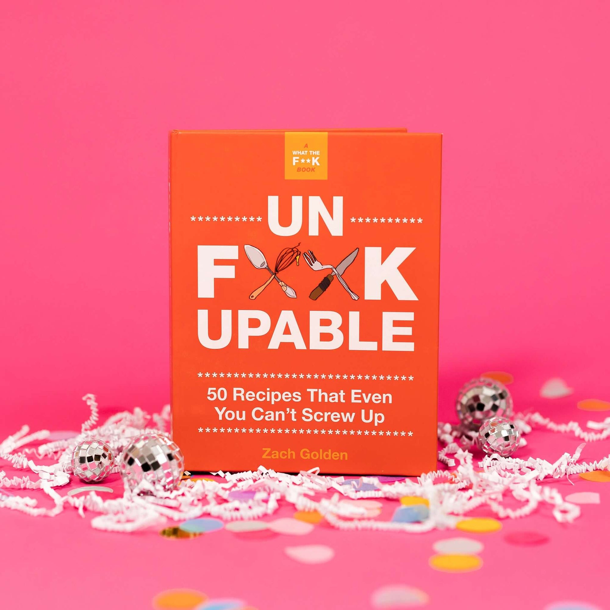 On a hot pink background sits a book with white crinkle and big, colorful confetti scattered around. There are mini disco balls. It is called "A WHAT THE F**K BOOK." It has illustraions of utensils crossed and in white it says "UN F**KUPABLE" in all caps, block font. It also says "50 Recipes that Even You Can't Screw Up" in white, block font. By Zach Golden.