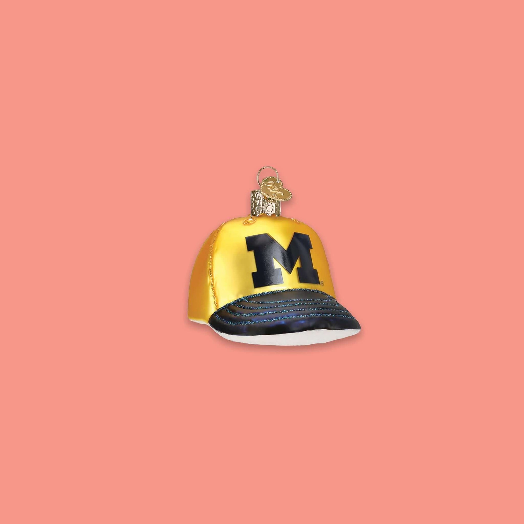 On a coral pink background sits a maize and navy University of Michigan ball cap glass ornament. It has the letter "M" on it.