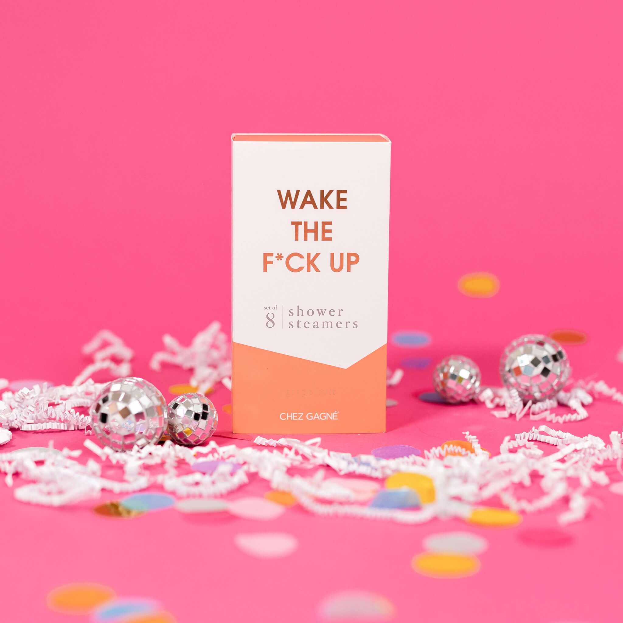 On a hot pink background sits a box with white crinkle and big, colorful confetti scattered around. There are mini disco balls. This picture is a close-up of a white and peach package that says "WAKE THE F*CK UP" in gold foil, all caps block lettering. Under it says "set of 8" and " shower steamers" in grey, lowercase serif font. At the bottom it says "grapefruit" in gold foil, lower case serif font.