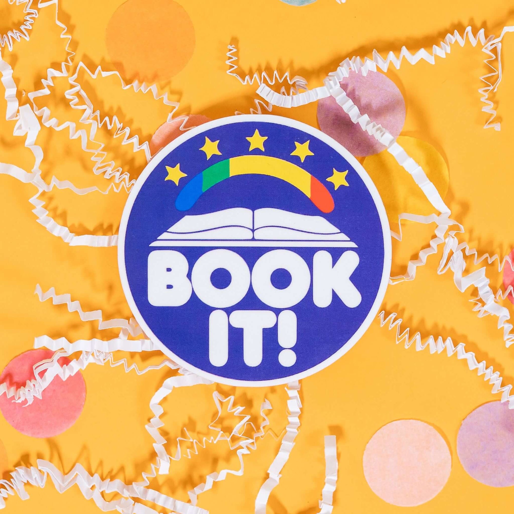 On a sunny mustard background sits a sticker with white crinkle and big, colorful confetti scattered around. This brilliant blue purple round sticker has a white illustration of an open book a rainbow and 5 yellow stars above it. It says "BOOK IT!" in a white, bubble text.