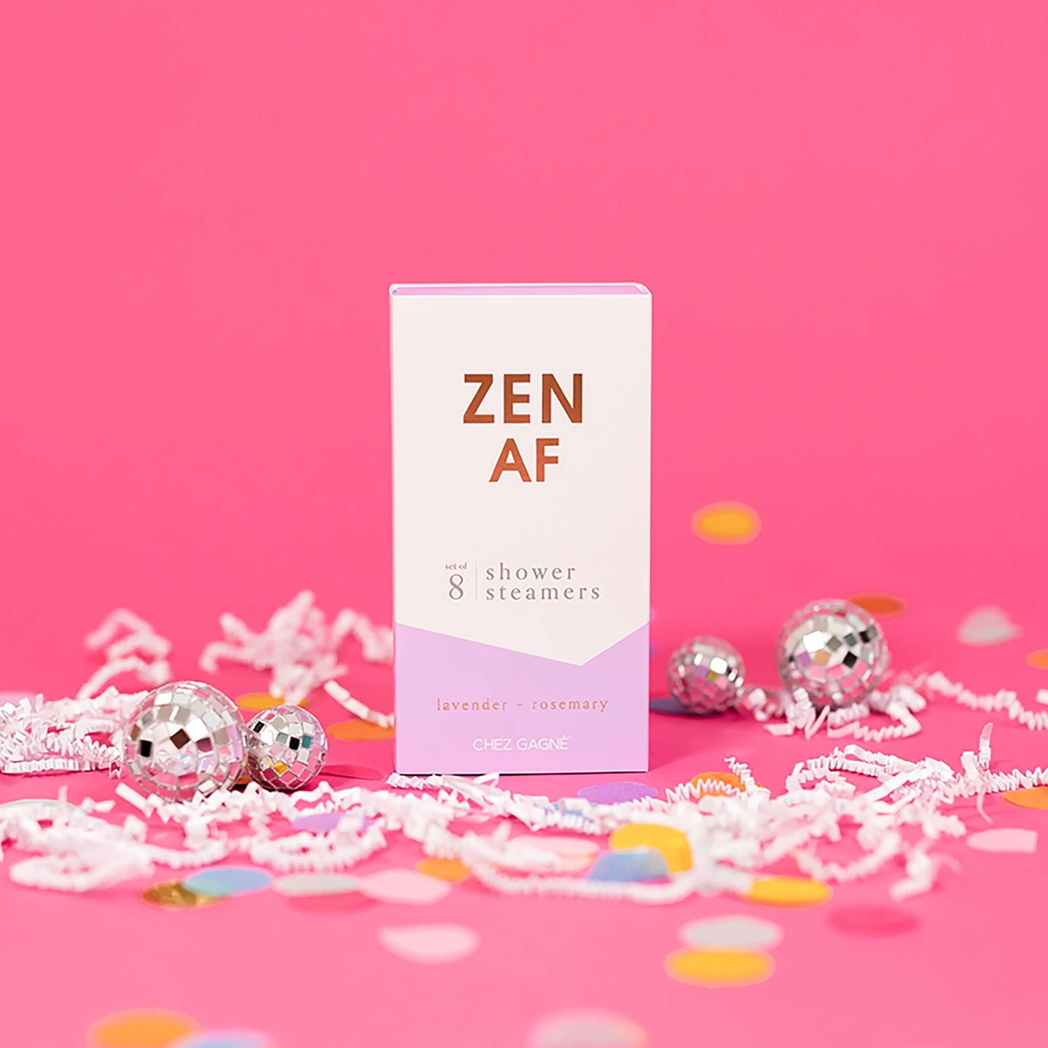 On a hot pink background sits a box with white crinkle and big, colorful confetti scattered around. There are mini disco balls. This picture is a close-up of a white and lavender package that says "ZEN AF" in gold foil, all caps block lettering. Under it says "set of 8" and " shower steamers" in grey, lowercase serif font. At the bottom it says "lavender - rosemary" in gold foil, lower case serif font. To the right of it is a lilac box with lavender shower steamers in it.