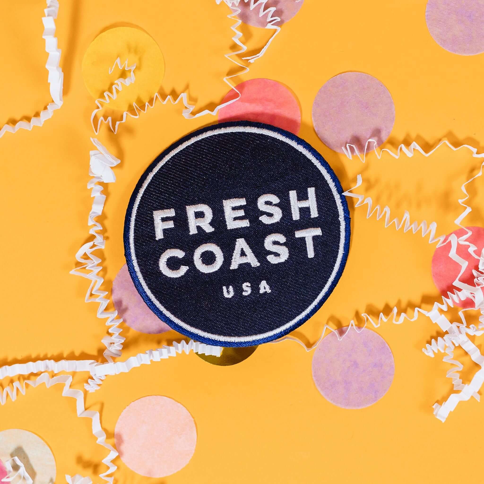 On a sunny mustard background sits a patch with white crinkle and big, colorful confetti scattered around. This navy patch has a white outline circle stitching on the edge and in the center in white it says "FRESH COAST USA" in block stitch lettering.