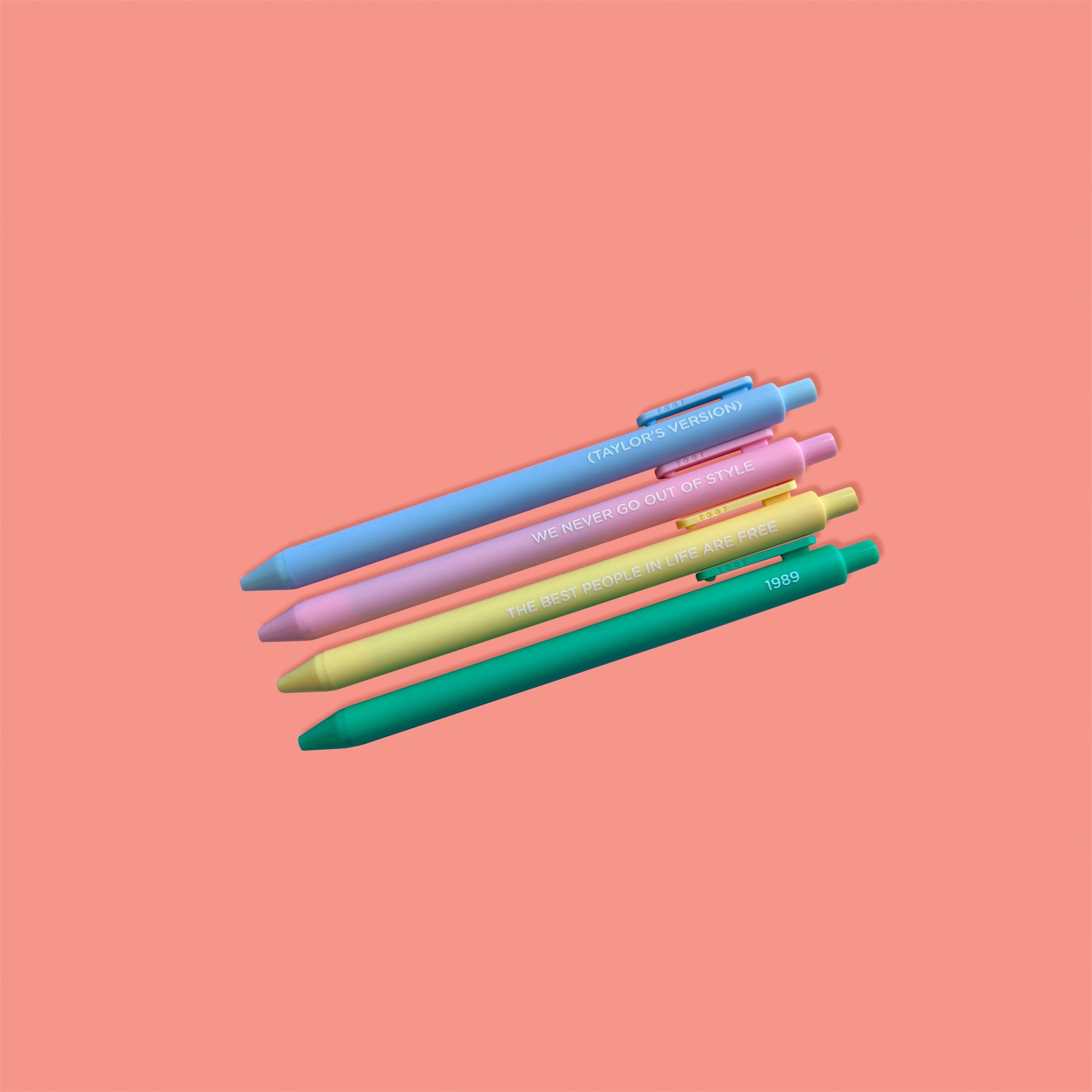 On a coral pink background sits a set of 4 pens. These Taylor Swift inspired pens are in pastel colors of blue, pink, yellow, and green. They say "(TAYLOR'S VERSION)", "WE NEVER GO OUT OF STYLE", "THE BEST PEOPLE IN LIFE ARE FREE", AND "1989." 