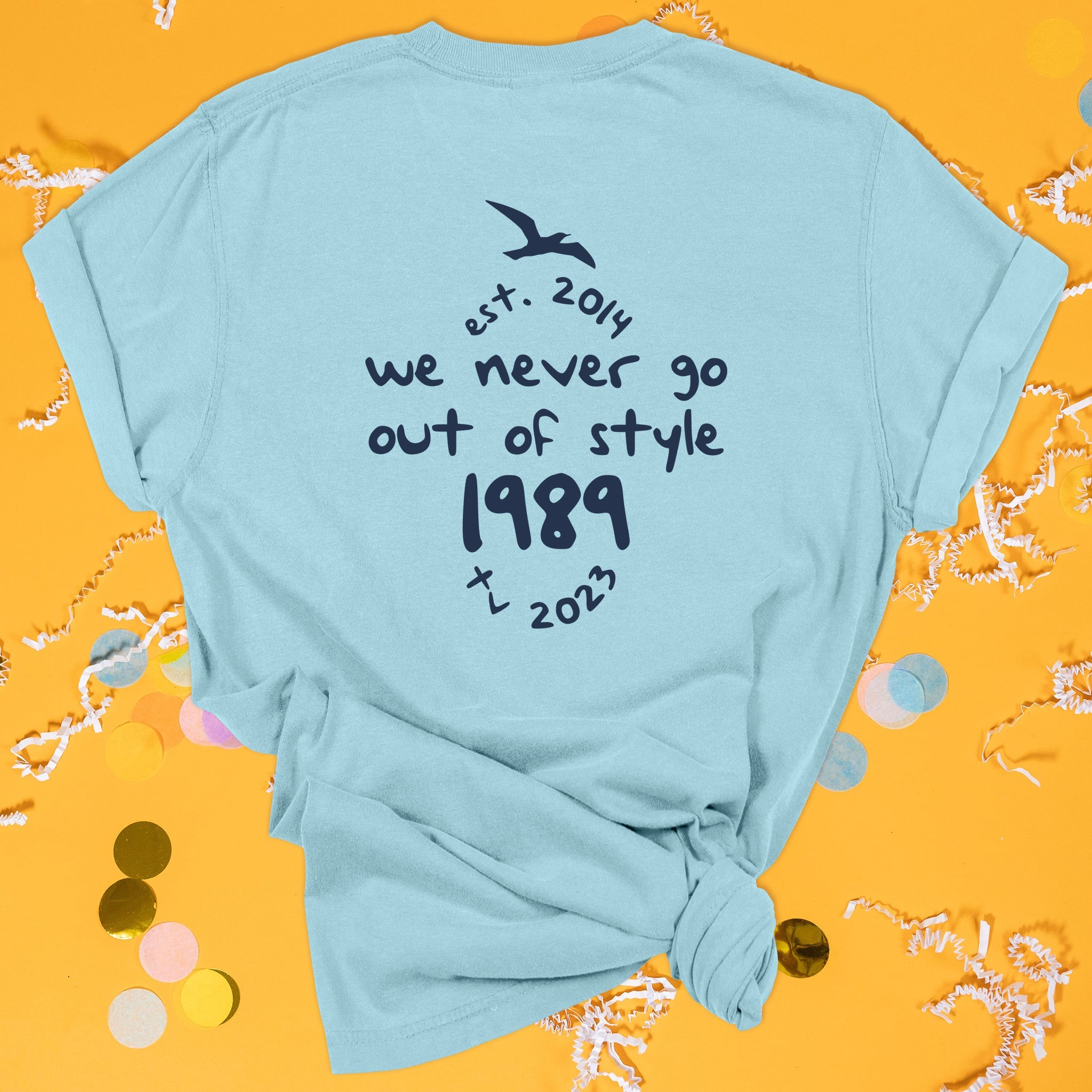 On a sunny mustard background sits the back of a t-shirt with white crinkle and big, colorful confetti scattered around. This Taylor Swift Inspired Reputation tee is aqua with navy lettering and an illustration of a seagull. It says "we never go out of style 1989" and "est. 2014 tv 2023" in handwritten lettering.