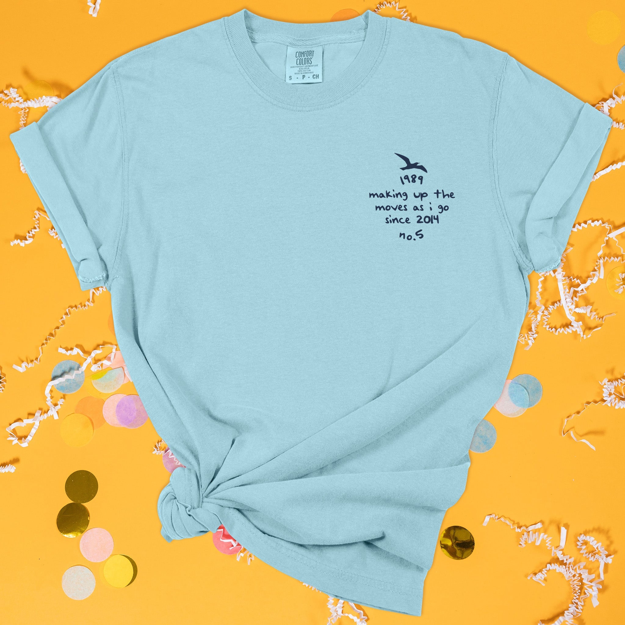 On a sunny mustard background sits the front of a t-shirt with white crinkle and big, colorful confetti scattered around. This Taylor Swift Inspired Reputation tee is aqua with navy lettering and an illustration of a seagull over the left chest area. It says "making up the moves as I go since 2014" and "1989 no.5" in handwritten lettering.