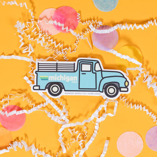 On a sunny mustard background sits a sticker with white crinkle and big, colorful confetti scattered around. This white sticker has an illustration of a light blue truck with black outlines and it says "michigan USA" in white block lettering with three stripes. The stripes colors are yellow, green, and blue. 2"-3"