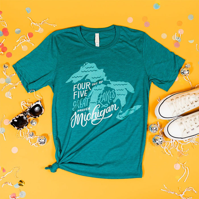 On a sunny mustard background sits a t-shirt with white crinkle and big, colorful confetti scattered around. There are mini disco balls, sunglasses, and a pair of white sneakers. The teal t-shirt features an illustration of Lakes Erie, Huron, Michigan, and Superior boring the great Mitten State with hand lettering. It is in white and aquamarine. 