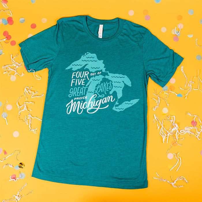 On a sunny mustard background sits a t-shirt with white crinkle and big, colorful confetti scattered around. The teal t-shirt features an illustration of Lakes Erie, Huron, Michigan, and Superior boring the great Mitten State with hand lettering. It is in white and aquamarine. 