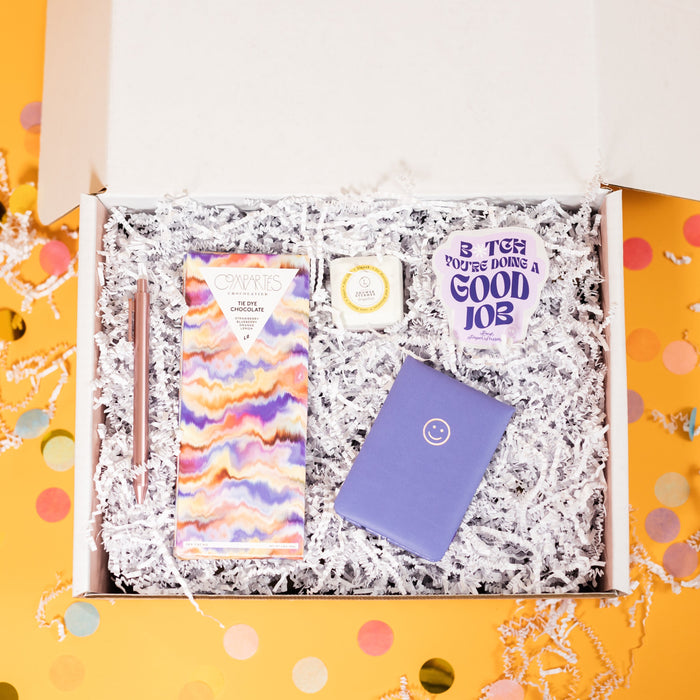 On a sunny mustard background sits a box pretty pastel gifts on a bed of white crinkle. There are big, colorful confetti surrounding the box. The Smiley Face Notepad is purple with a smiley face on the top. There is a lavender sticker that says "B*tch you're doing a good job" in a purple, groovy font with "Rock Paper Scissors" below it in a handwritten font and a happy face in white, a Rose Gold Pen, Compoartes Tie Dye Chocolate Bar, and Lizush Grapefruit Shower Steamer.