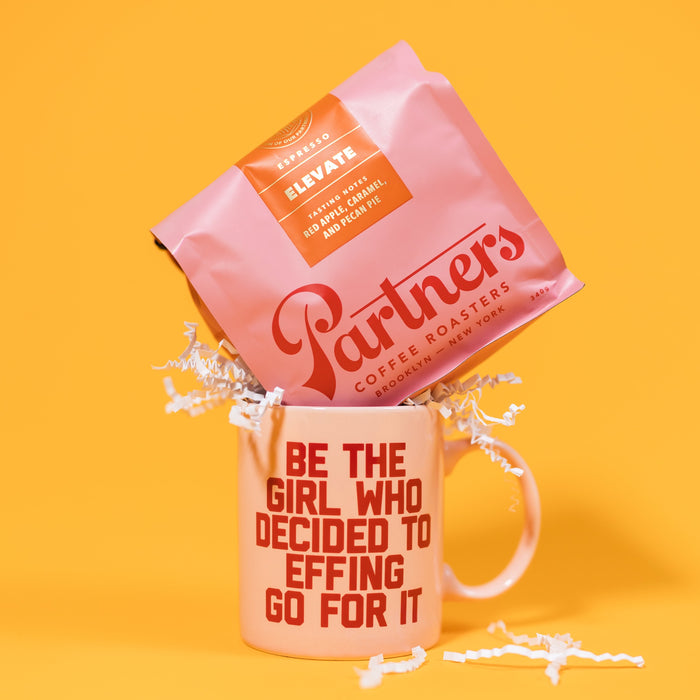 On a mustard yellow background sits a mug and a bag of coffee. The pink bag of Partners coffee sits atop the pink diner style mug with red capital collegiate writing that says "Be the girl who decided to effing go for it." White crinkle also overflows out of the mug.