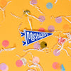On a sunny mustard background sits a magnet with white crinkle and big, colorful confetti scattered around. This 2D-style raised magnet is a blue and white handdrawn pennant that says "Michigan" in white handdrawn script lettering. 3"-4"