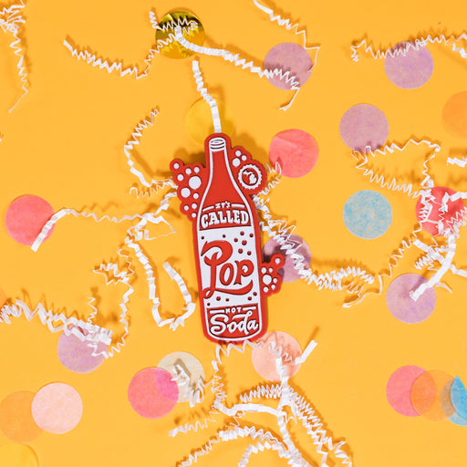 On a sunny mustard background sits a magnet with white crinkle and big, colorful confetti scattered around. This 2D-style raised magnet is a red and white handdrawn soda pop that says "It's Called Pop Not Soda" in red and white handwritten lettering. It has a white outline bubble with the state of Michigan in it in white. 3"- 4"