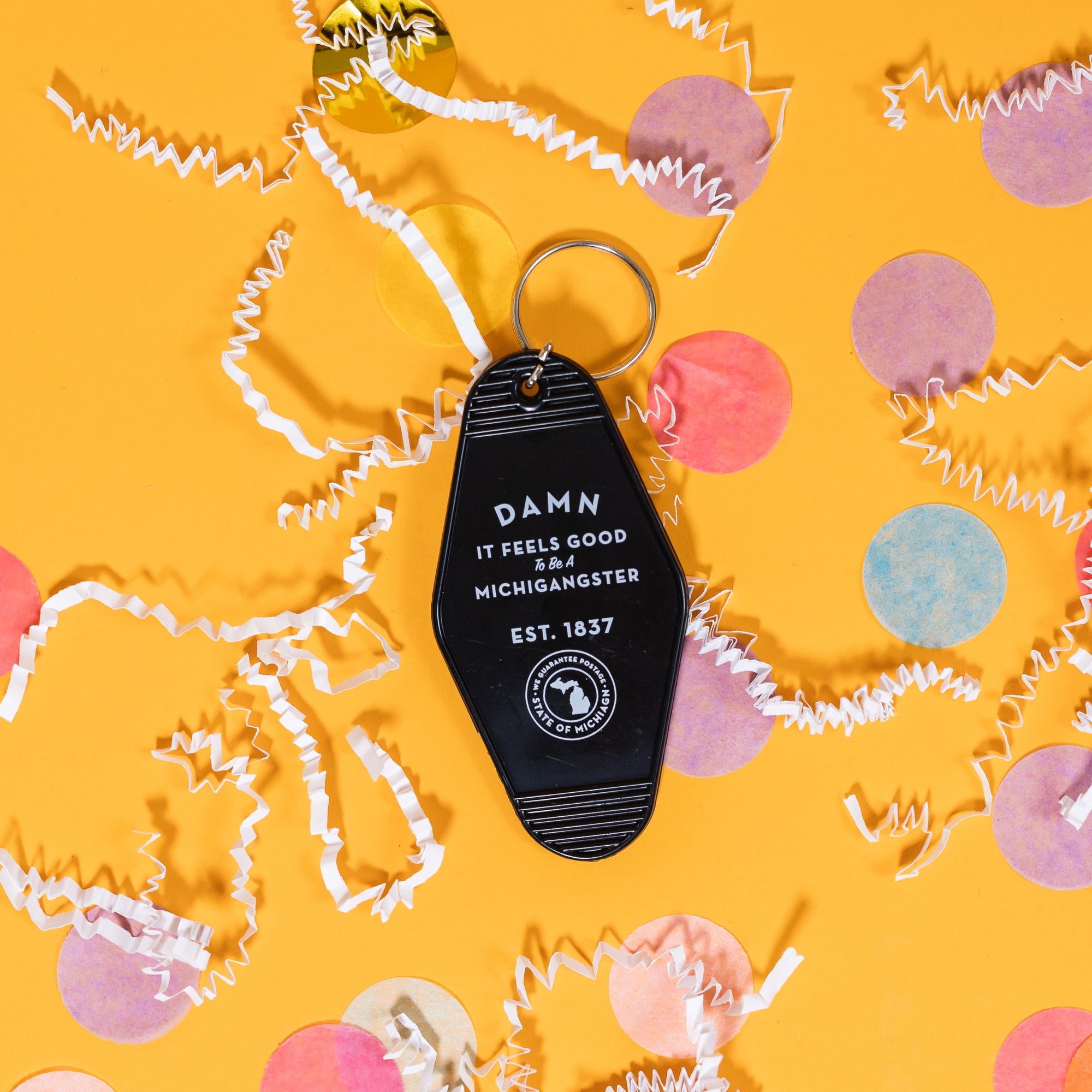 On a sunny mustard background sits a hotel key with white crinkle and big, colorful confetti scattered around. This black vintage hotel key ring says "Damn It Feels Good To Be A Michigangster EST. 1837" in white text. There is a design that includes an icon of Michigan's lower and upper peninsulas enclosed in a cirle that reads "State of Michigan" in white.