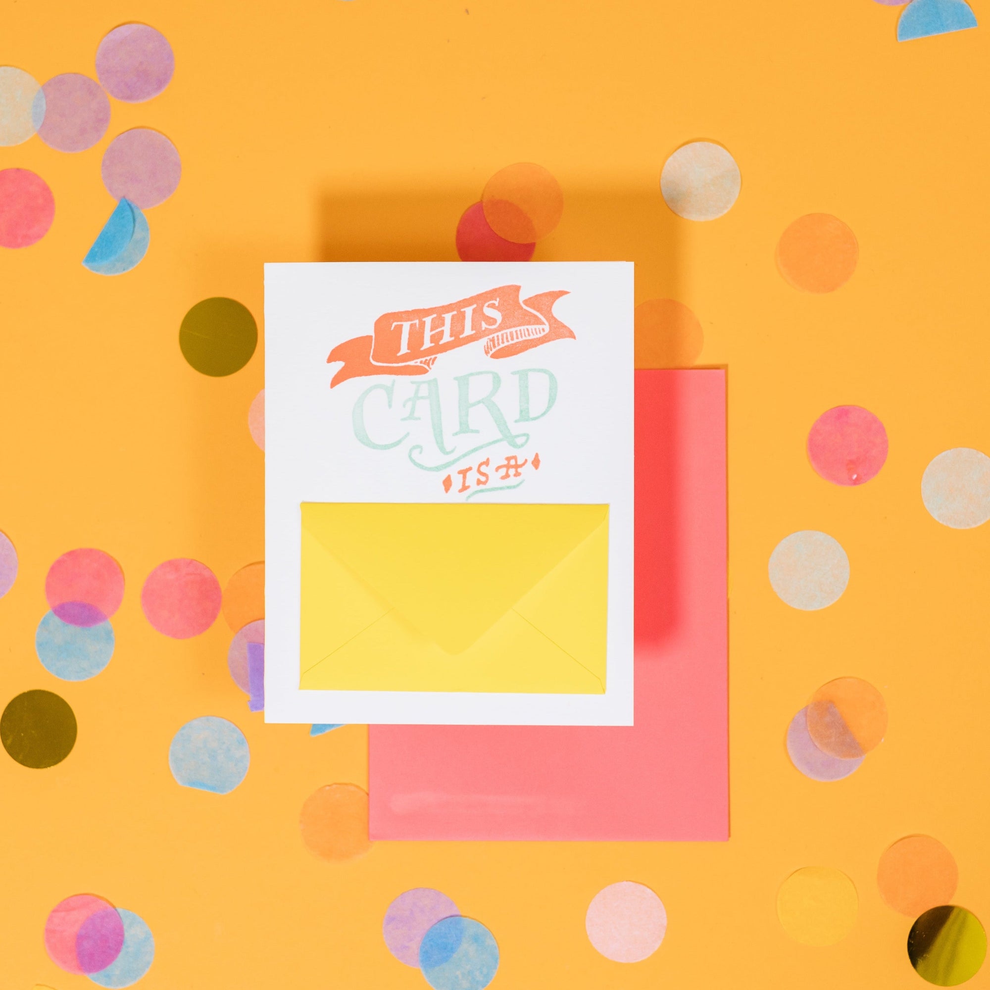 On a sunny mustard background is a greeting card and envelope with big, colorful confetti scattered around. The white greeting card says "This Card Is A" in handwritten vintage orange and cool aqua letterpress lettering. There is a white mini card inside small yellow envelope on the front that says "HUG" in orange in a big, bold font. The watermelon pink envelope sits under the card. 
