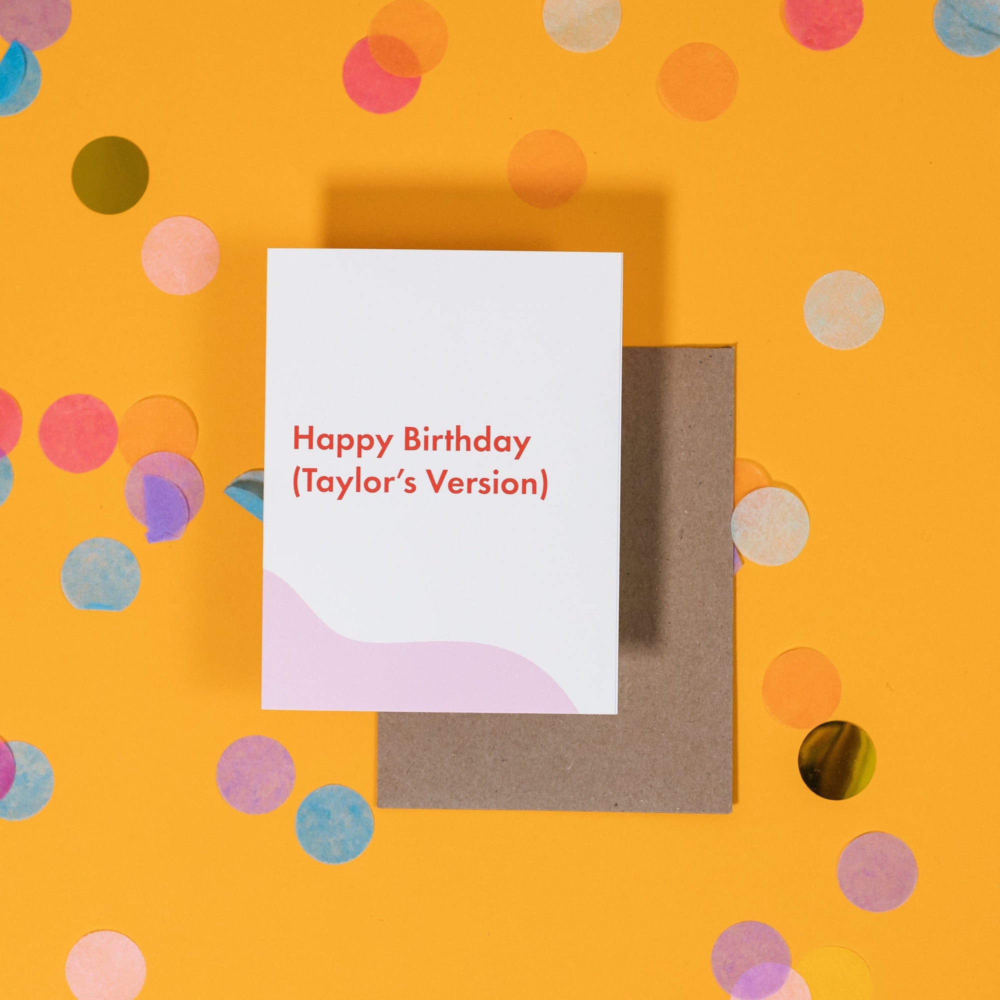 On a sunny mustard background is a greeting card and envelope with big, colorful confetti scattered around. The white greeting card says "Happy Birthday (Taylor's Version)" in sans serif red font. The neon red envelope sits under the card. 