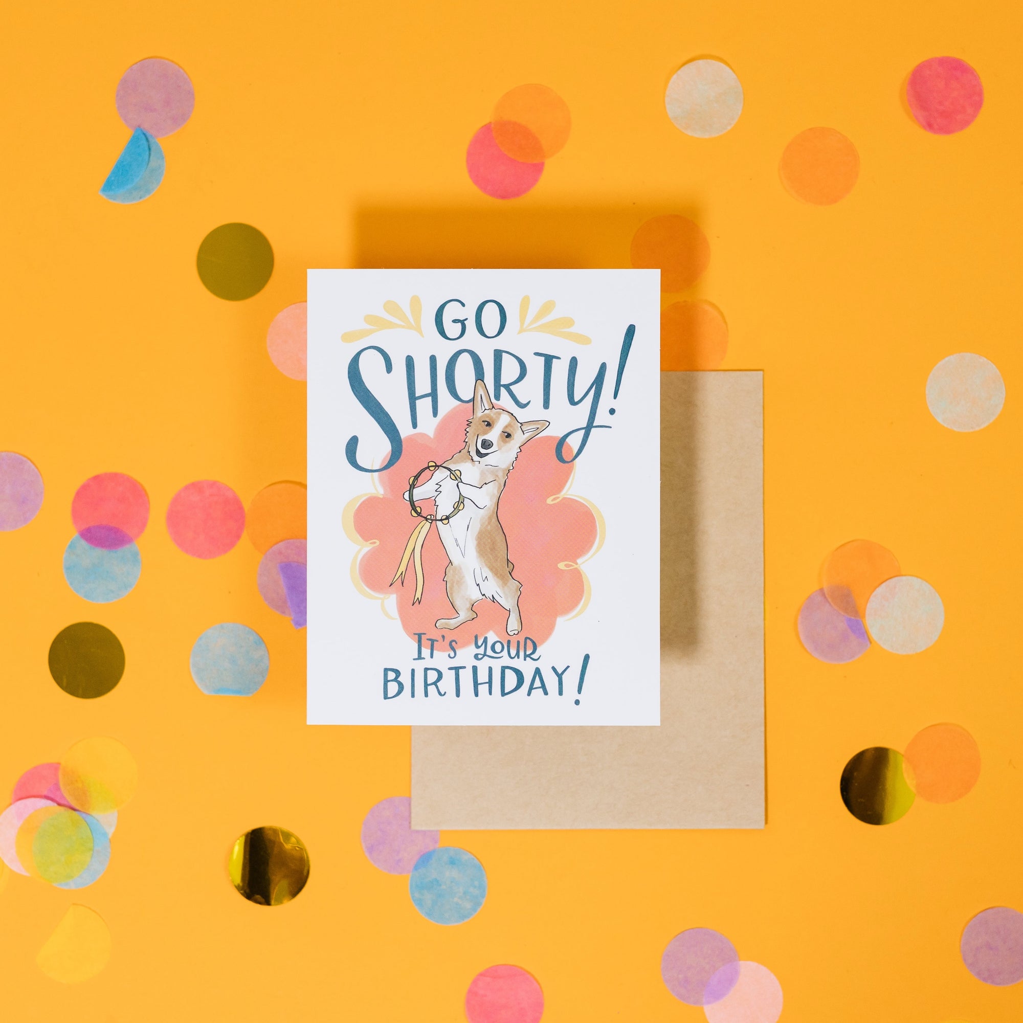 On a sunny mustard background is a greeting card and envelope with big, colorful confetti scattered around. The white greeting card has an illustration of a Corgi dog playing the tamborine on it and says "Go Shorty It's Your Birthday!" in handwritten blue lettering. The kraft envelope sits under the card. 