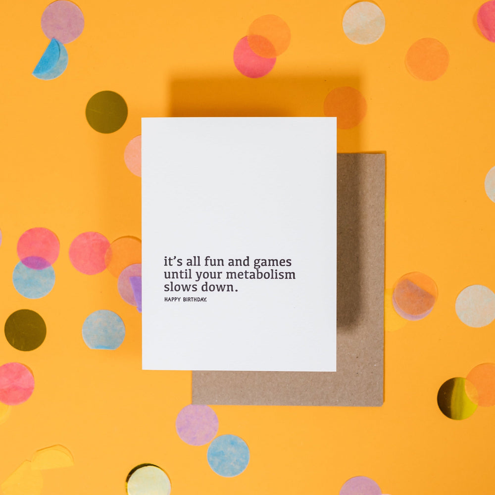 On a sunny mustard background is a greeting card and envelope with big, colorful confetti scattered around. The white greeting card  says "it's all fun and games until your metabolism slows down." in a black, lowercase serif font. It also says "HAPPY BIRTHDAY" in a black, all caps block font. A kraft envelope sits under the card.