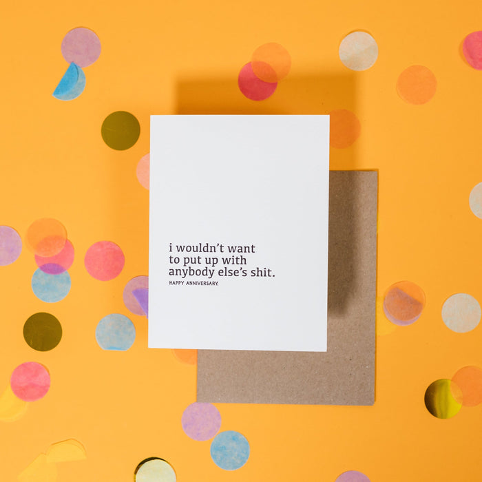 On a sunny mustard background is a greeting card and envelope with big, colorful confetti scattered around. The white greeting card says "I wouldn't want to put up with anybody else's shit. Happy Anniversary" in all lowercase serif black letterpress text. The kraft envelope sits under the card. 