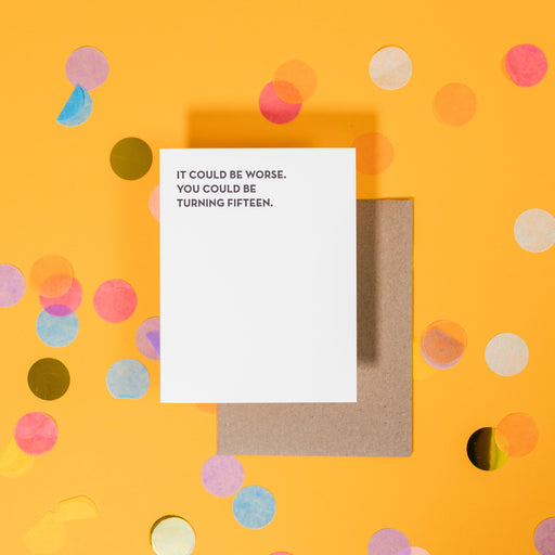 On a sunny mustard background is a greeting card and envelope with big, colorful confetti scattered around. The white greeting card says "It Could Be Worse. You Could Be Turning Fifteen." in a black, sans serif uppercase font. The kraft envelope sits under the card. 