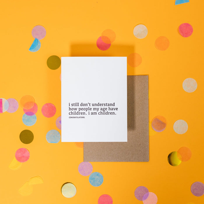 On a sunny mustard background is a greeting card and envelope with big, colorful confetti scattered around. The white greeting card says "I still don't understand how people my age have children. I am children. congratulations." in a black, lower case serif font. A kraft envelope sits under the card.