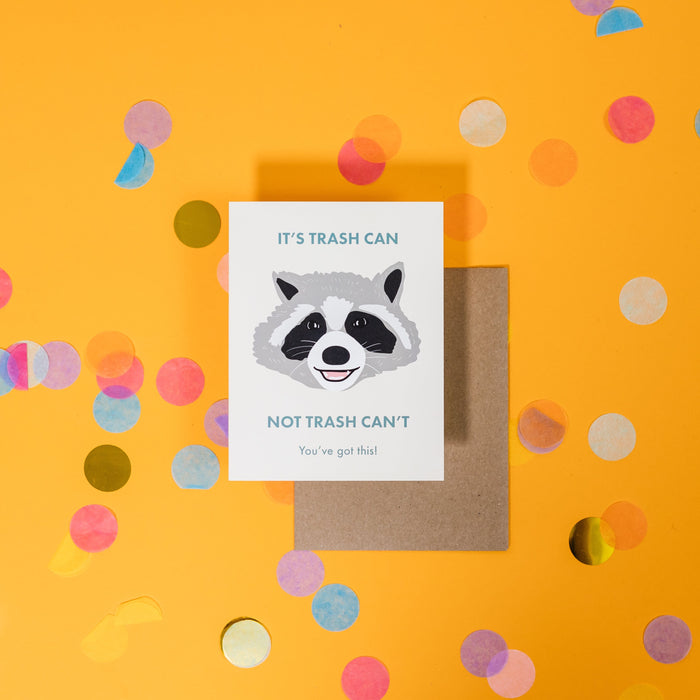 On a sunny mustard background is a greeting card and envelope with big, colorful confetti scattered around. The white greeting card has an illustration of a raccoon face and says "It's Trash Can Not Trash Can" in an aquamarine sans serif font. Under that says "You've got this!" is smaller, aquamarine font. The kraft envelope sits under the card. 