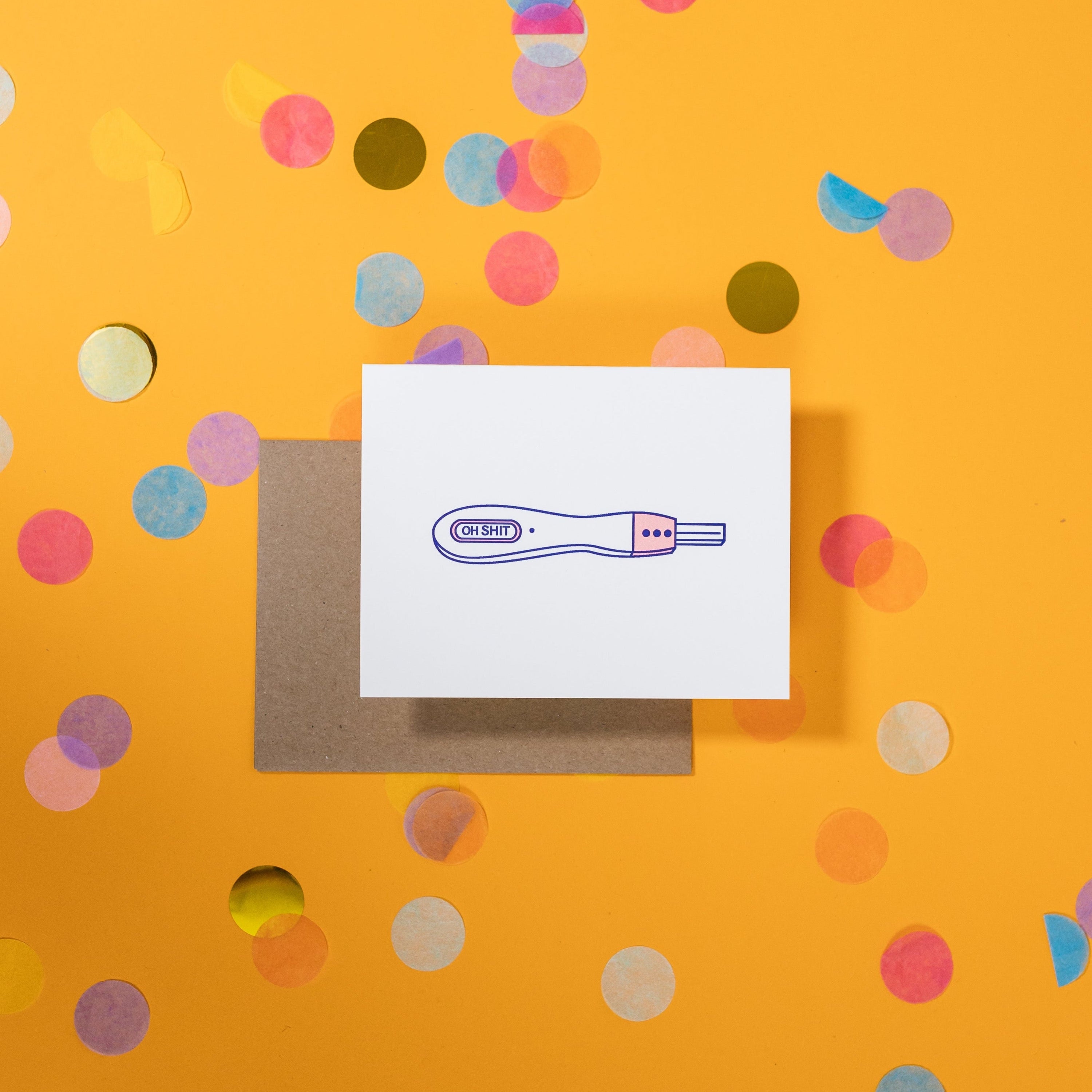 On a sunny mustard background is a greeting card and envelope with big, colorful confetti scattered around. The white greeting card has an illustration of a pregnancy test that says "OH SHIT" in a black, block letterpress text. The kraft envelope sits under the card. 