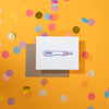 On a sunny mustard background is a greeting card and envelope with big, colorful confetti scattered around. The white greeting card has an illustration of a pregnancy test that says "OH SHIT" in a black, block letterpress text. The kraft envelope sits under the card. 