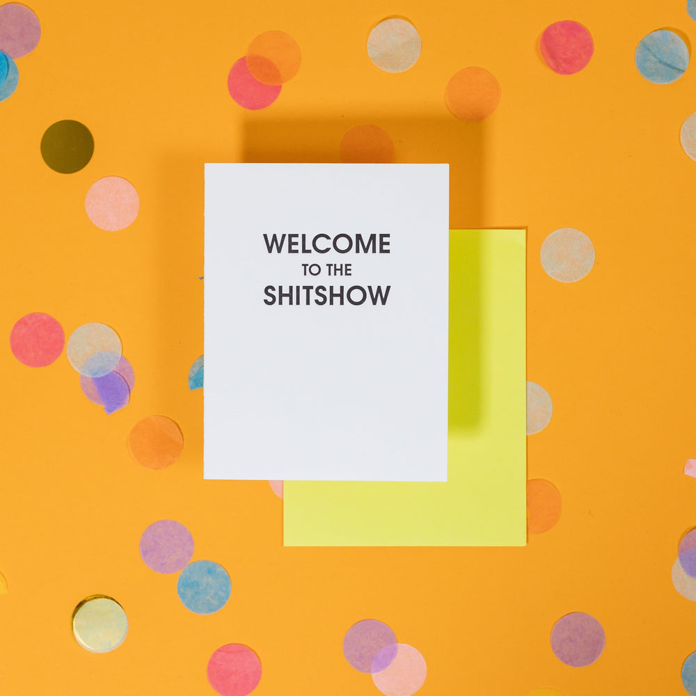 On a sunny mustard background is a greeting card and envelope with big, colorful confetti scattered around. The white greeting card says "WELCOME TO THE SHITSHOW" in big black, sans serif uppercase font. The neon yellow envelope sits under the card. 