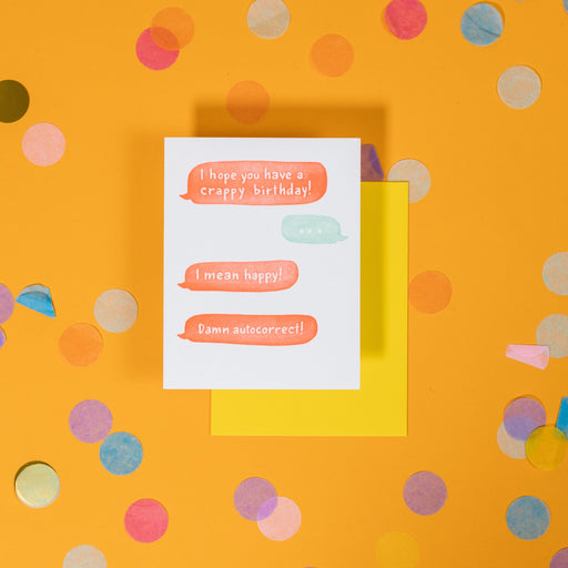 On a sunny mustard background is a greeting card and envelope with big, colorful confetti scattered around. The white greeting card has an illustration of text bubbles in orange and light blue that says "I hope you have a crappy birthday!", "...", "I mean happy!", "Damn autocorrect" in handwritten white font. The yellow envelope sits under the card. 