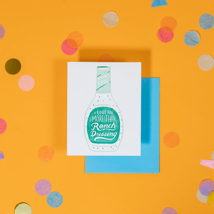 On a sunny mustard background is a greeting card and envelope with big, colorful confetti scattered around. The white greeting card has an illustration of a Ranch Dressing bottle in aqua green and says "100% True I Love You More Than Ranch Dressing" in handwritten white lettering. The turquoise blue envelope sits under the card. 