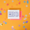 On a sunny mustard background is a greeting card and envelope with big, colorful confetti scattered around. The white greeting card says "HBD" in handwritten funky bold lettering in light blue and navy. The neon red envelope sits under the card. 