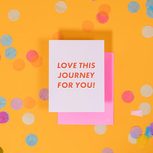 On a sunny mustard background is a greeting card and envelope with big, colorful confetti scattered around. The light pink greeting card says "LOVE THIS JOURNEY FOR YOU!" in a block, italic red font with white drop shadow. The hot pink envelope sits under the card. 